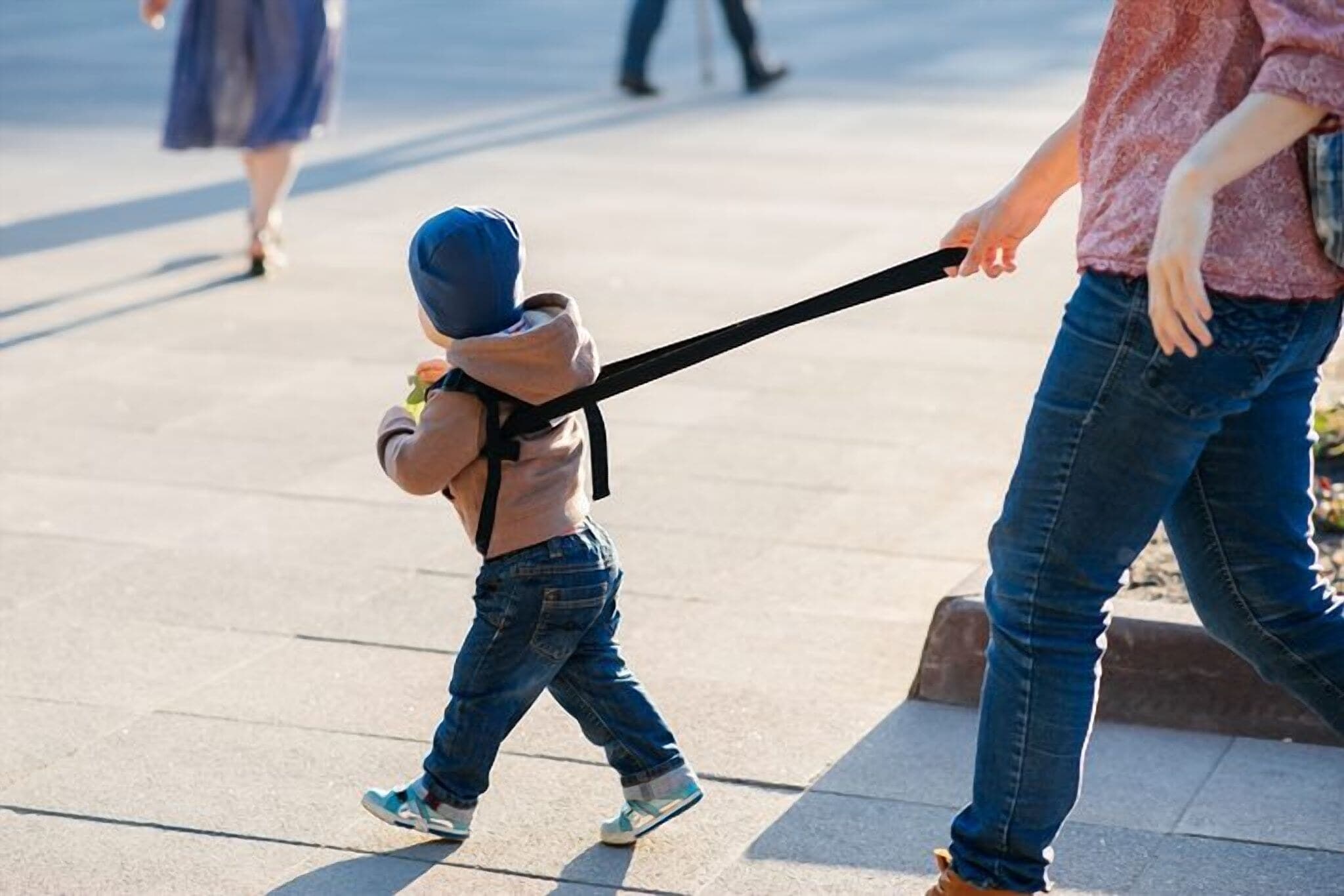 A parent holding a toddler by a safety harness strap while they walk along a city sidewalk